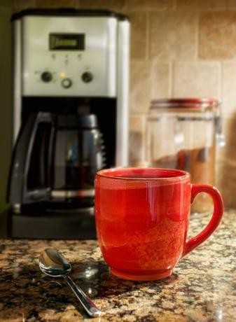 Naparovanie šálky kávy: Červený hrnček naparenej kávy spočíva na žulovej kuchynskej doske. Kávovar a nádoba na mletú kávu sú viditeľné na pozadí.